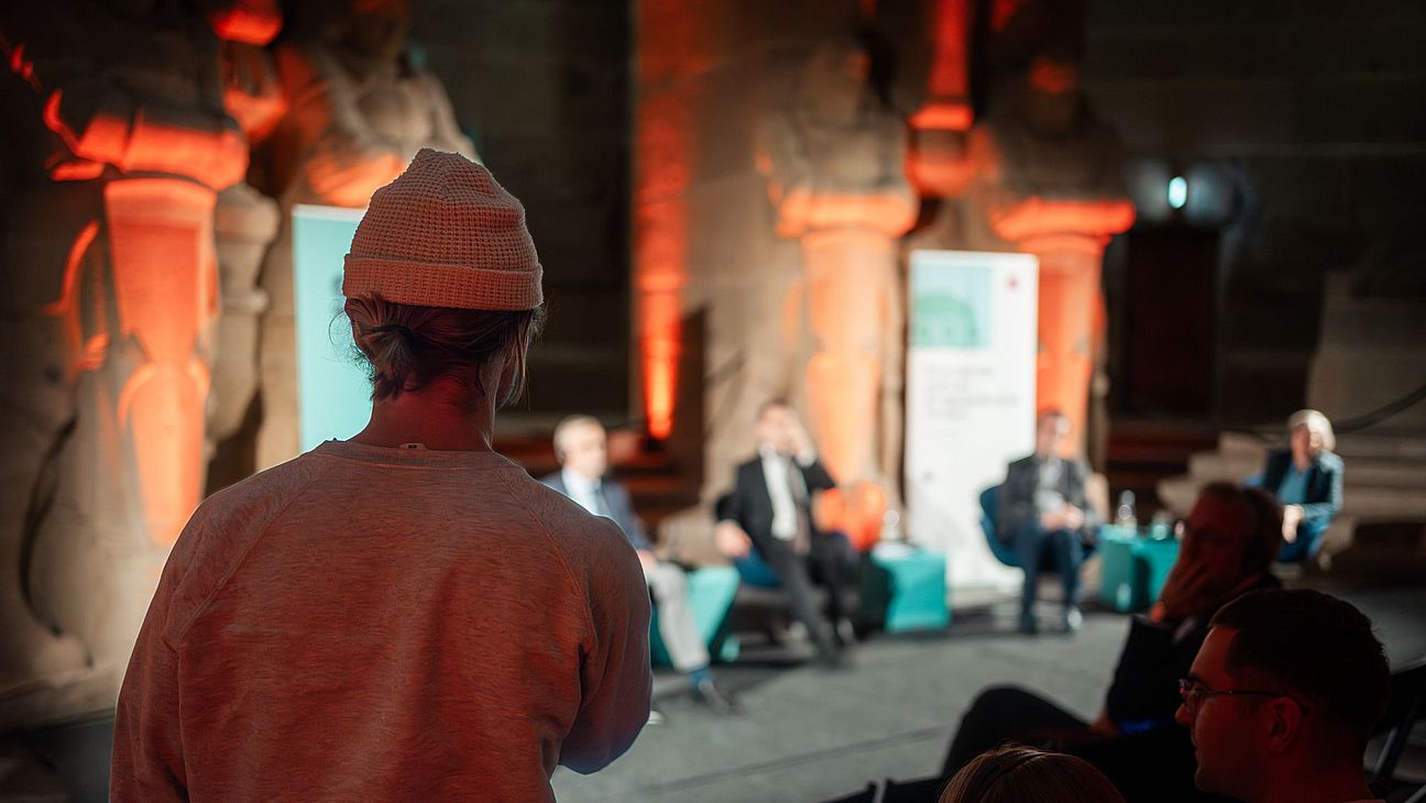 Person mit beiger Mütze und grauem Pullover steht im Vordergrund vor einer Podiumsdiskussion im Leipziger Völkerschlachtdenkmal mit orange beleuchteten Statuen im Hintergrund.