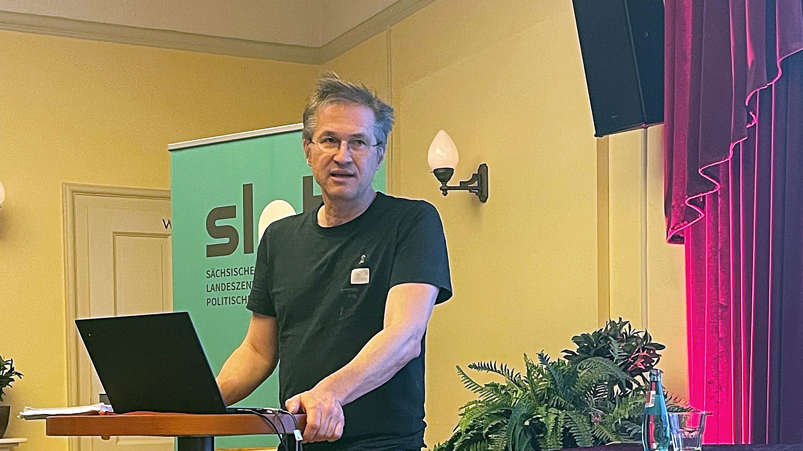 Mann in schwarzem T-Shirt steht an einem Stehpult mit Laptop in einem Raum mit Holzboden und rotem Vorhang, im Hintergrund Banner mit Schriftzug 'slpb'