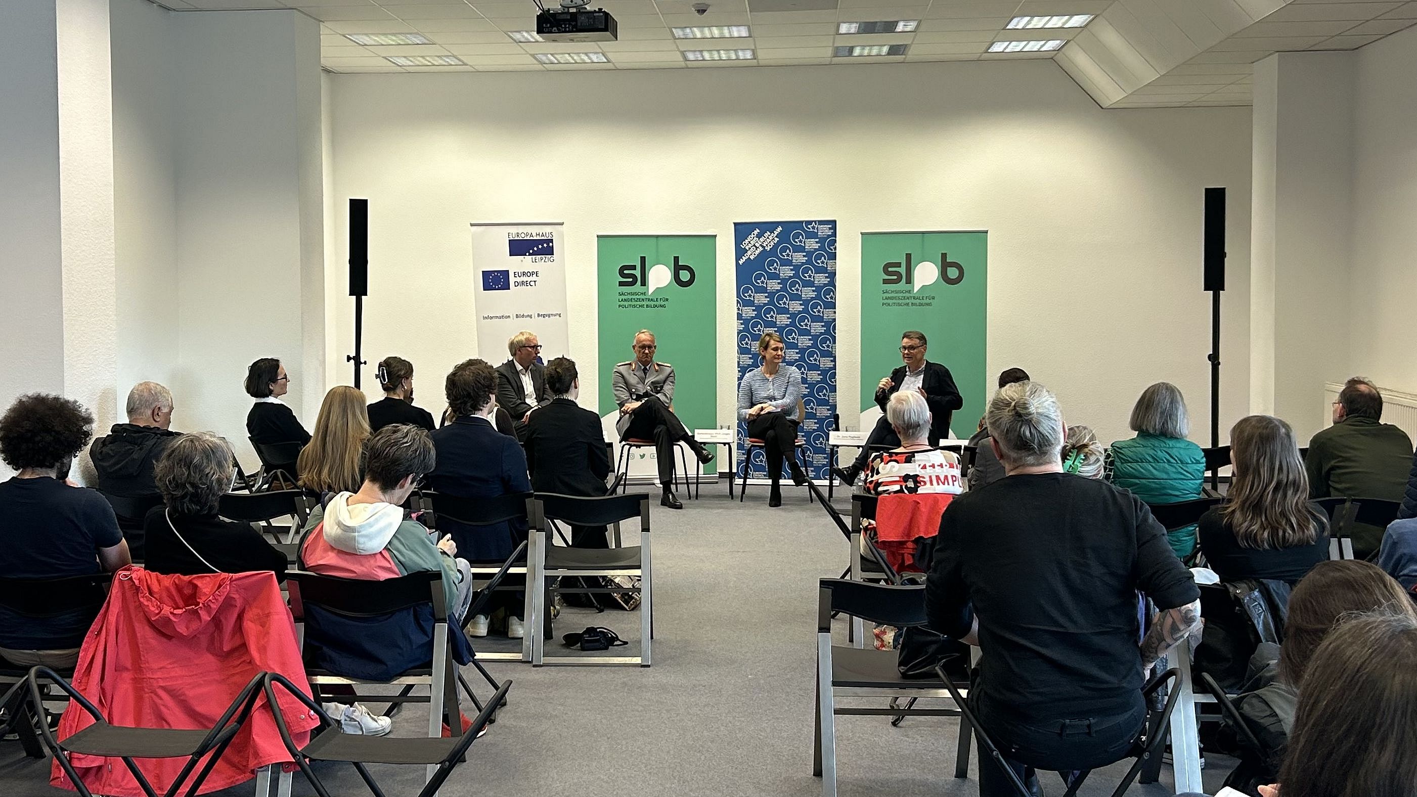 Im Vordergrund sind auf der linken und rechten Seite die Teilnehmerinnen der Veranstaltung auf ihren Stühlen sitzend zu sehen. Im Hintergrund sitzt das Podium.
