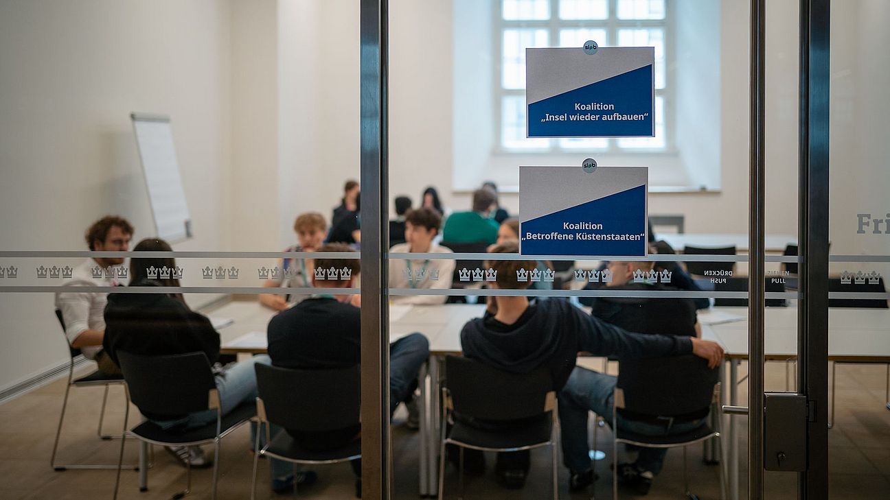 Hinter einer Glaswand sitzen junge Menschen an Konferenztischen.