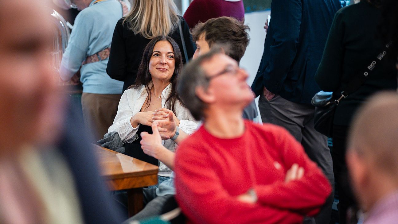 Mehrere Personen stehen und sitzen in einem Raum, einige unterhalten sich. Eine Frau mit langem Haar trägt eine weiße Bluse und sitzt an einem Holztisch.