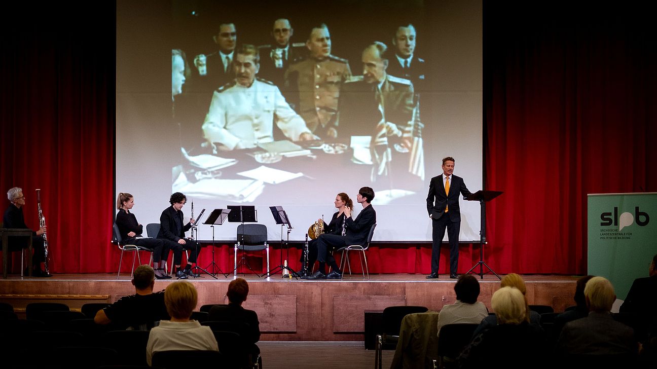Sechs Personen auf einer Bühne vor rotem Vorhang, fünf sitzen mit Musikinstrumenten, eine steht und hält ein Buch, im Hintergrund eine Projektion mit fünf Personen in historischen Uniformen.
