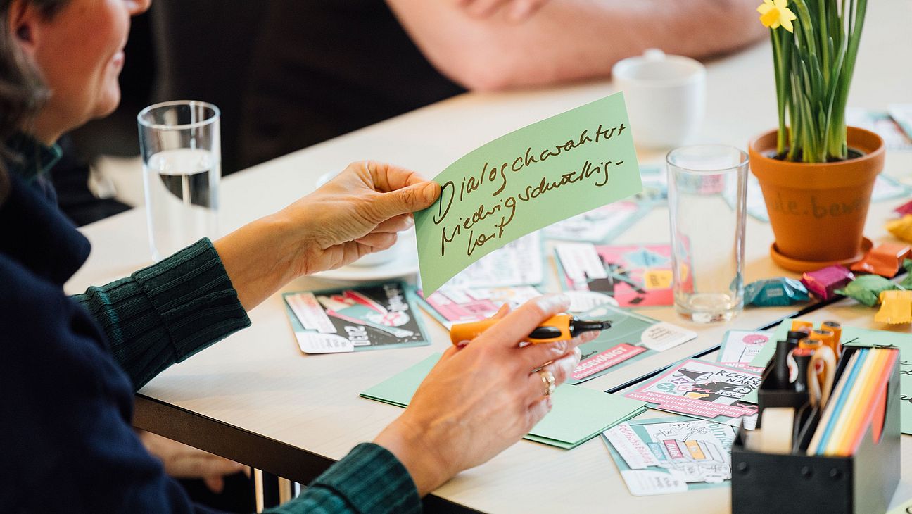KilligBild Person hält eine grüne Karte mit handschriftlichem Text 'Dialogcharakter, Niedrigschwelligkeit' über einem Tisch mit weiteren Karten, Stiften, Gläsern und einer Topfpflanze.