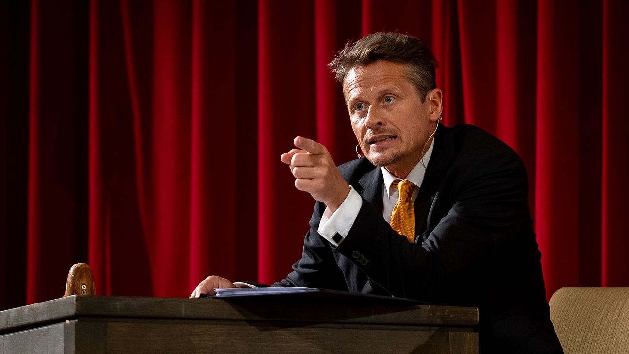 Mann in schwarzem Anzug mit weißem Hemd und gelber Krawatte sitzt an einem Holztisch vor rotem Vorhang und zeigt mit der rechten Hand nach vorne.