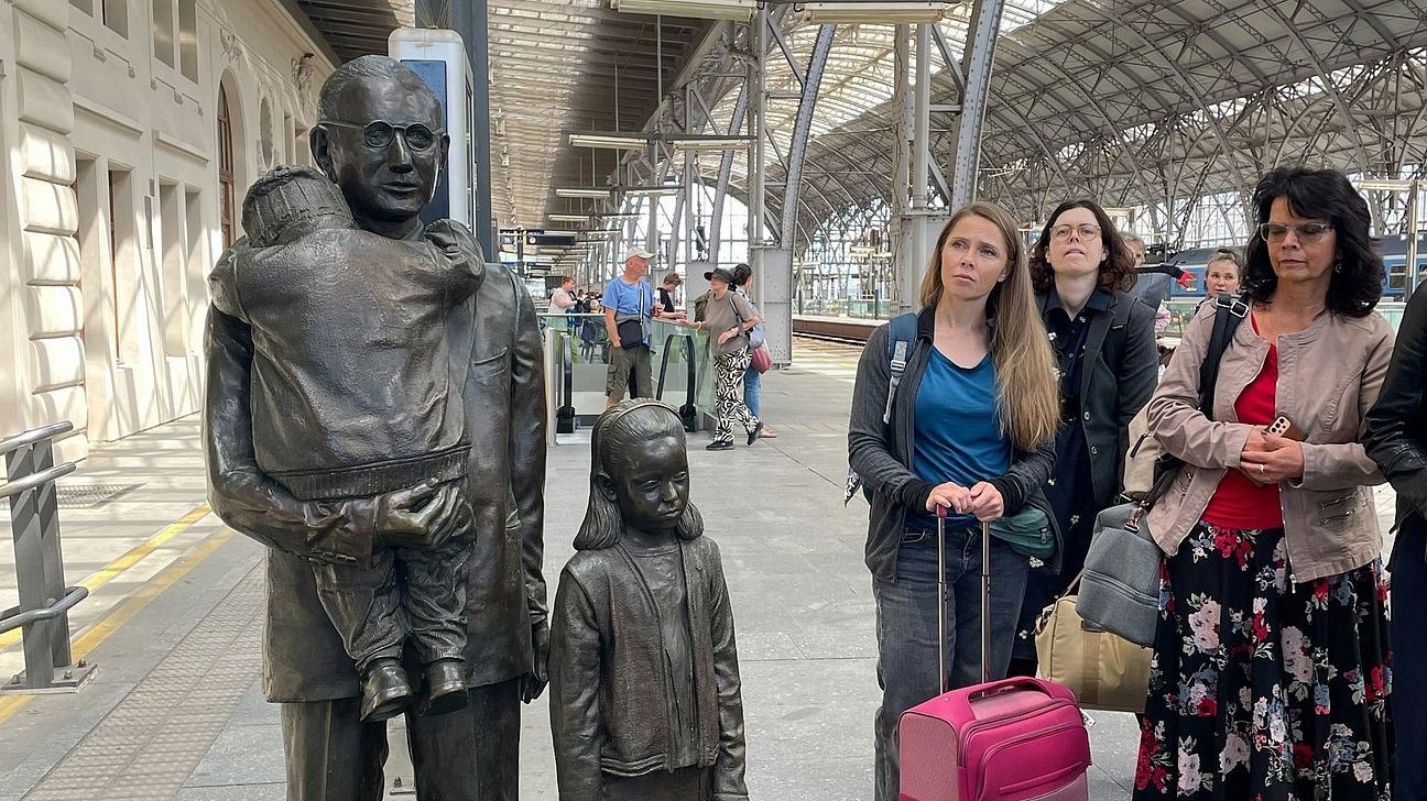 Im Vordergrund steht eine Statue mit einem kleinen Junge, der sich verzweifelt an den Hals eines älteren Mannes klammert, ein Mädchen steht mit leerem Blick daneben. Daneben eine Personengruppe die die Statue betrachtet.