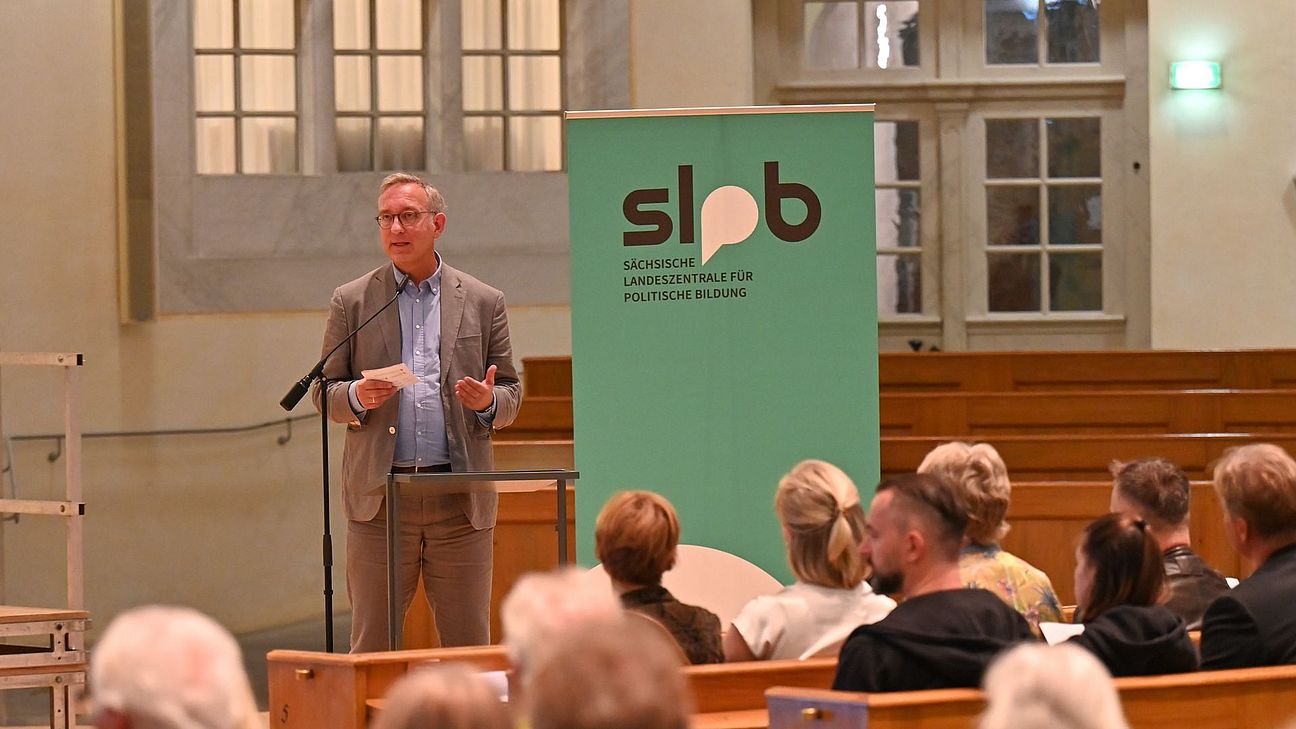 Ein Mann steht mit Mikrofon und Notizzettel vor einem Banner der SLpB und spricht zu einem Publikum in der Frauenkirche Dresden.