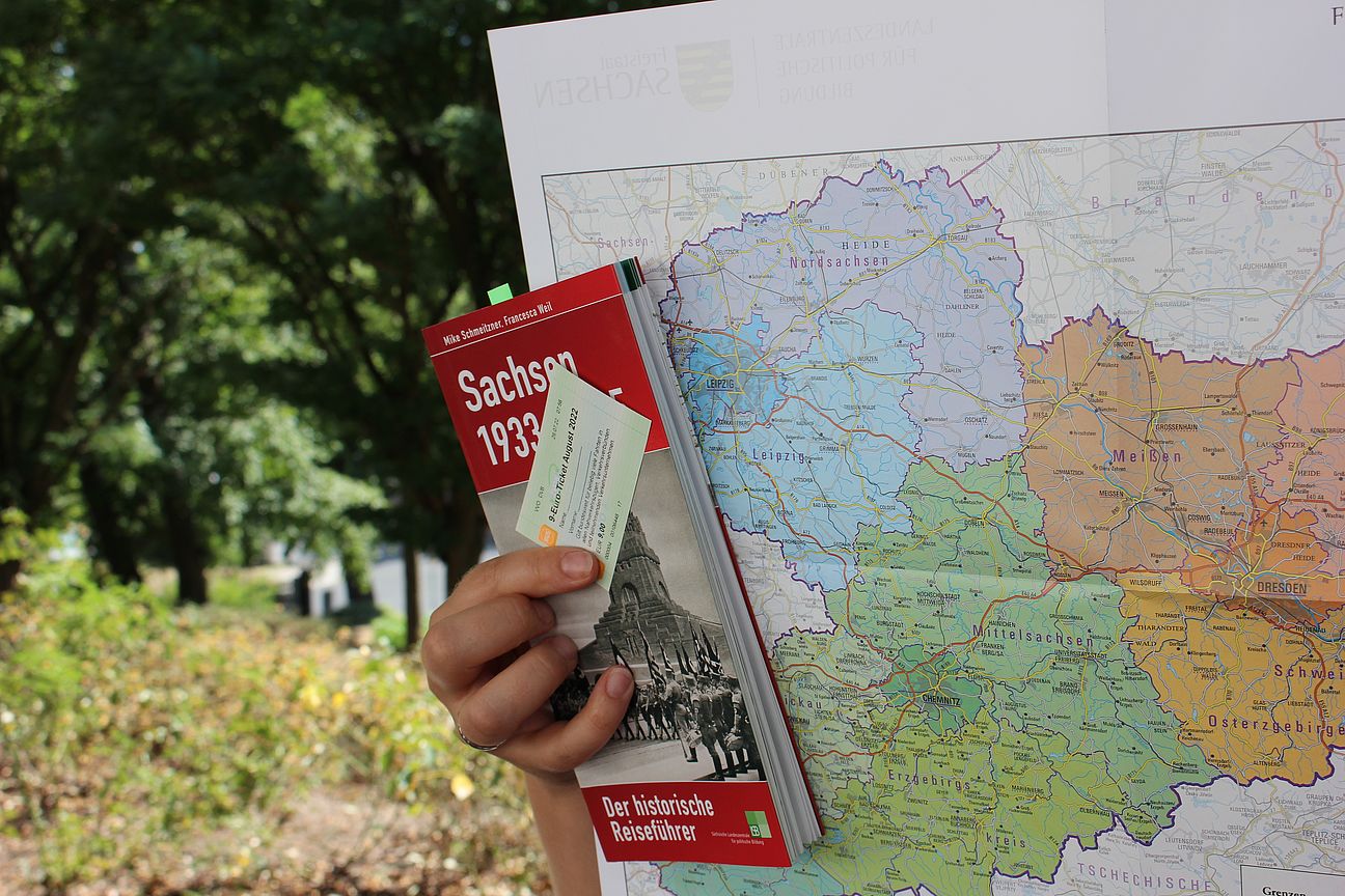 Reiseführer Symbolbild Zu sehen ist ein Reiseführer mit dem Titel "Sachsen 1933-1945" von Nora Schumann und Franziska Weil. Das Buch wird verlegt von der "Landeszentrale für politische Bildung Sachsen". Im Hintergrund ist eine Landkarte von Sachsen zu erkennen. Ein Zugticket mit der Aufschrift "DB Fahrkarte" wird ebenfalls in der Hand gehalten.