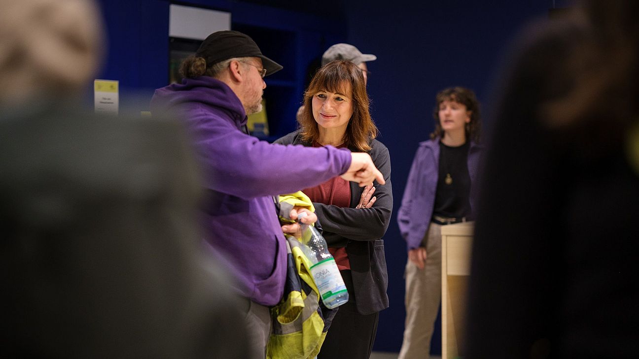 Mehrere Personen in einem Innenraum, eine Person trägt eine lila Kapuzenjacke und hält eine Flasche, andere Personen stehen im Hintergrund.