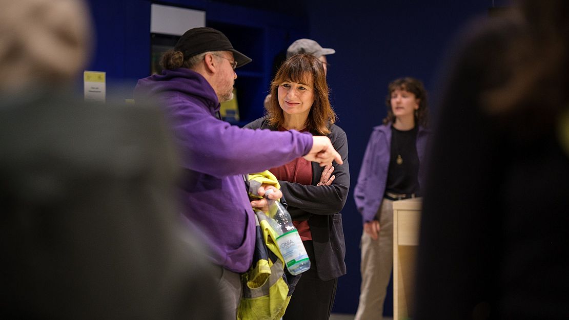 Mehrere Personen in einem Innenraum, eine Person trägt eine lila Kapuzenjacke und hält eine Flasche, andere Personen stehen im Hintergrund.