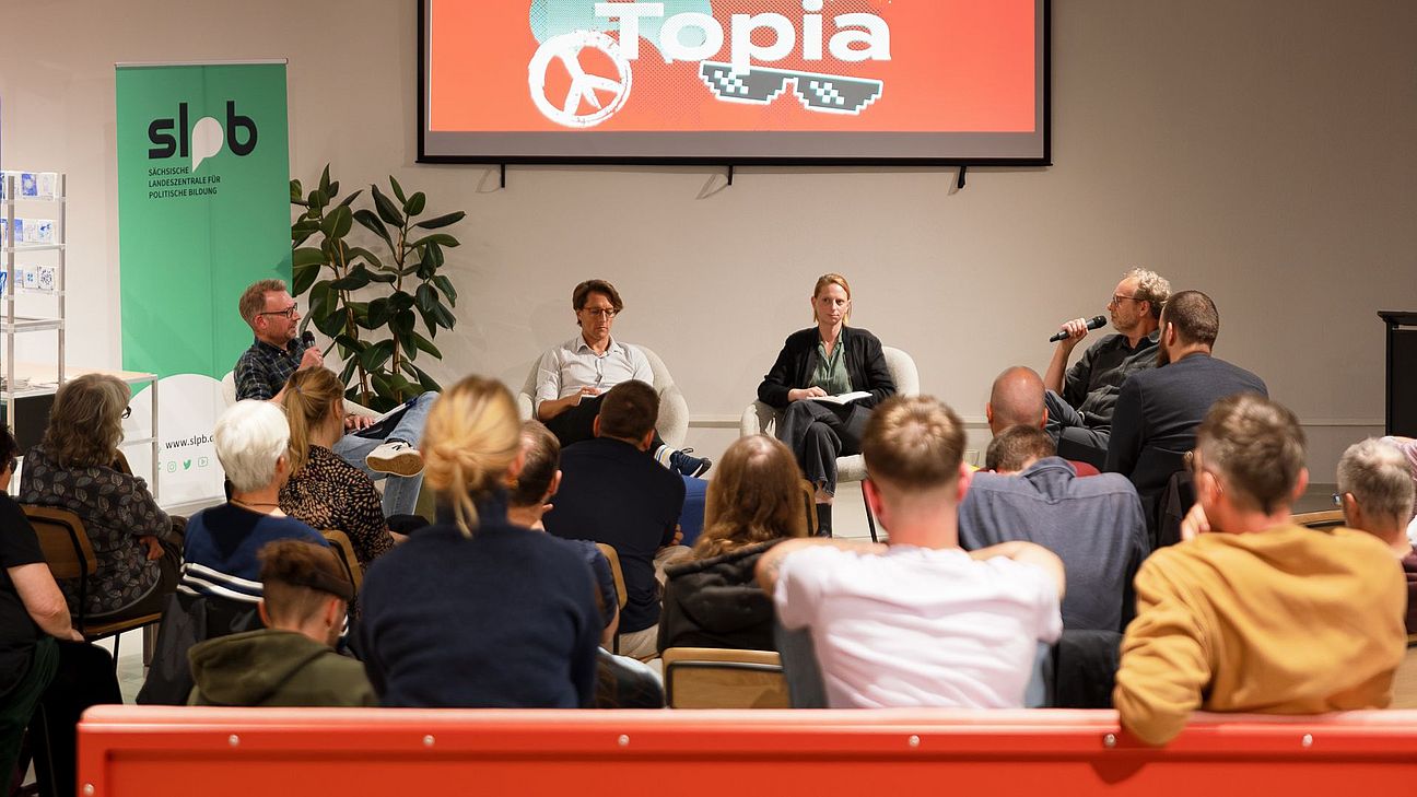 Auf einer Bühne diskutieren vier Personen vor einem Publikum. Im Hintergrund ist eine Präsentationsfolie mit dem Titel „Tech Bro Utopia“ sowie ein Banner der Sächsischen Landeszentrale für politische Bildung zu sehen.