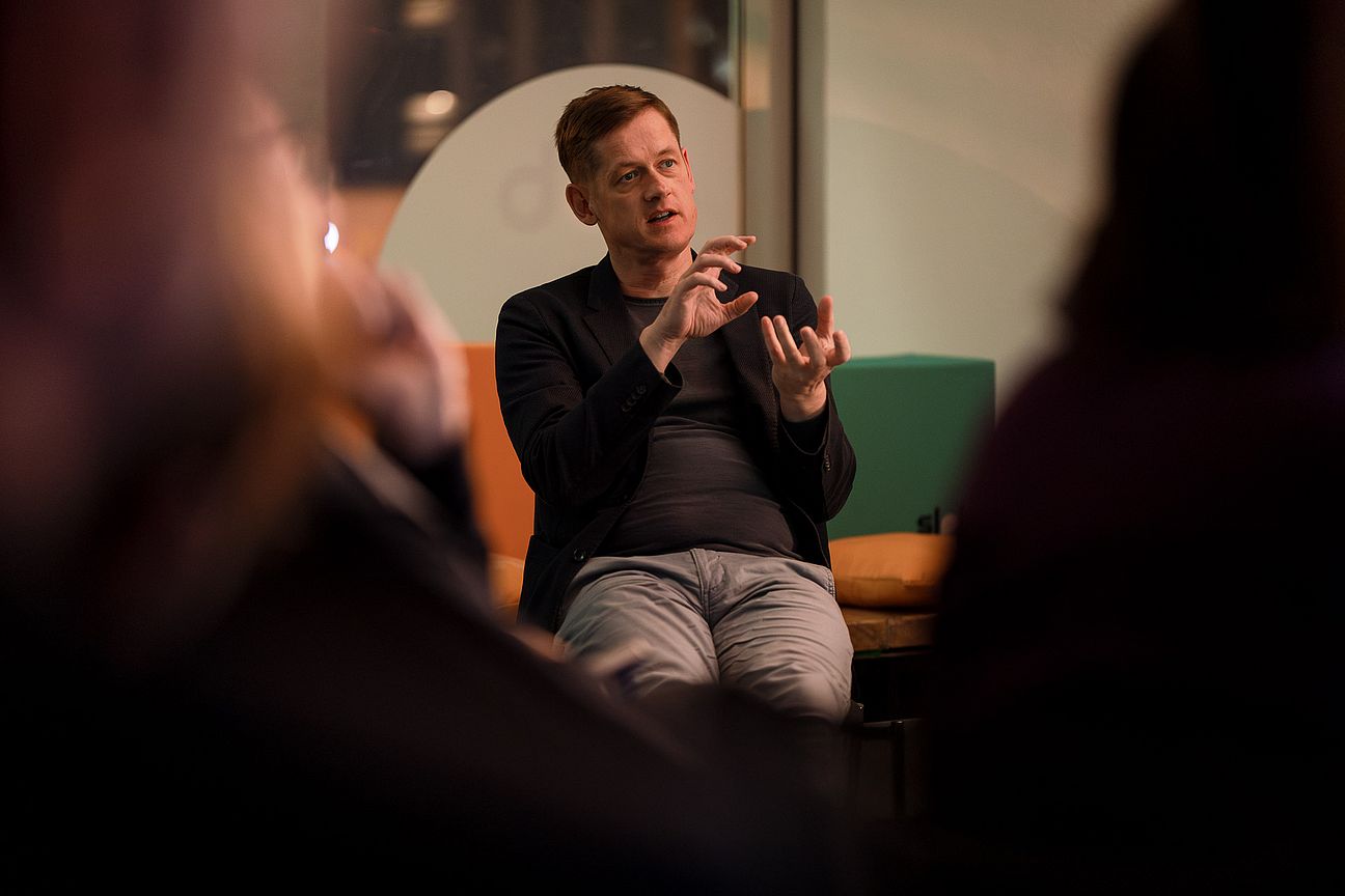 Mann sitzt auf Stuhl mit dem Rücken zu einem großen Fenster, im Vordergrund unscharfe Menschen aus dem Publikum.