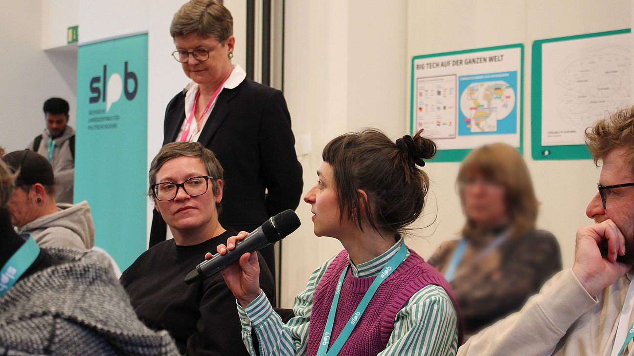 Frau mit Hochsteckfrisur und lila Strickweste hält Mikrofon, umgeben von mehreren sitzenden Personen in einem Raum mit SLpB-Banner und Informationsplakaten.