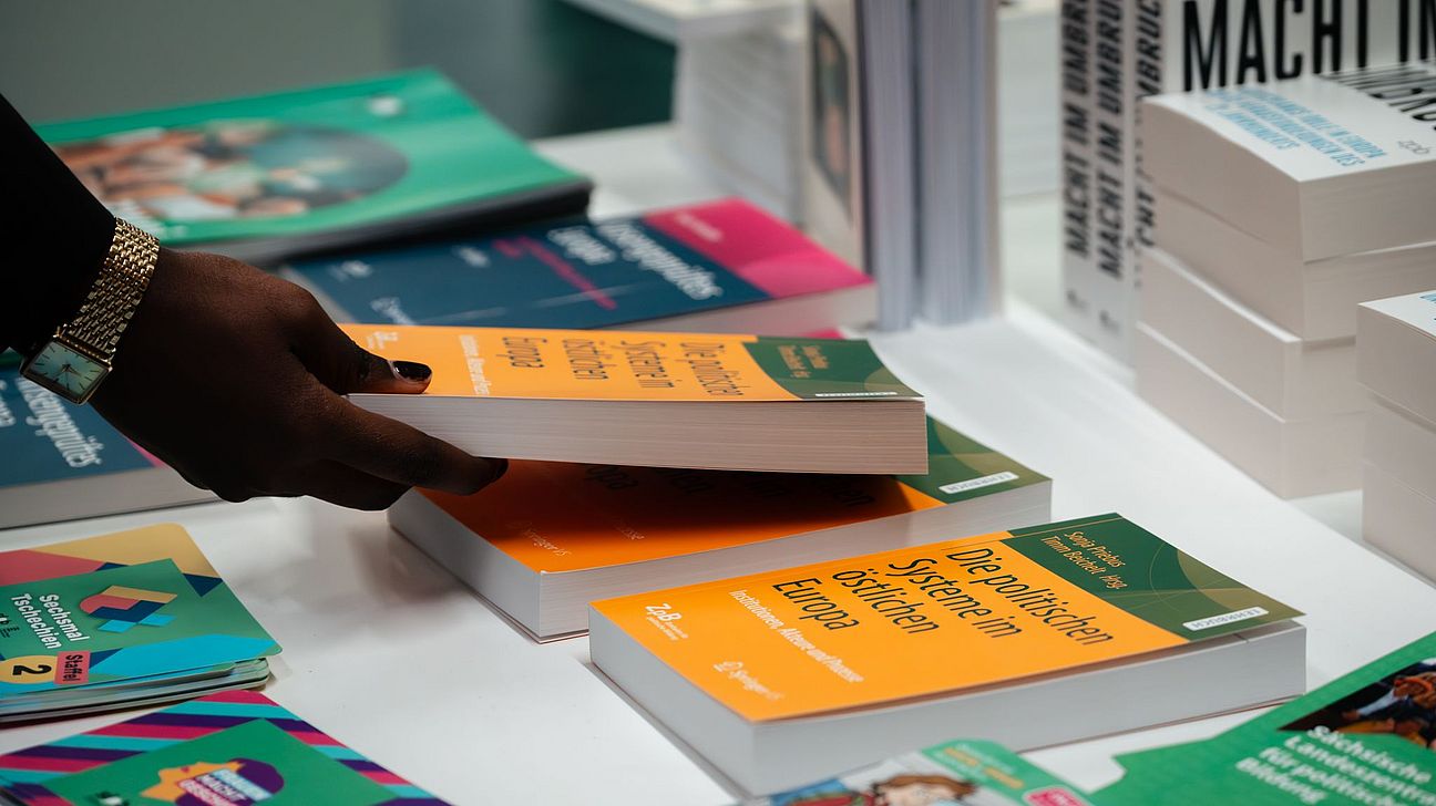 Hand mit goldfarbenem Armband nimmt ein orange-grünes Buch vom Büchertisch bei der Trinationalen Tagung, weitere Bücher liegen auf dem Tisch.