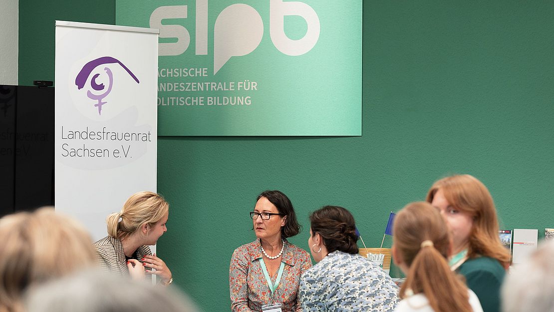 Drei Frauen sitzen am Tisch, eine spricht, während die anderen zuhören. Im Hintergrund steht ein Banner mit dem Text 'Landesfrauenrat Sachsen e. V.' und dem Logo der SLpB.