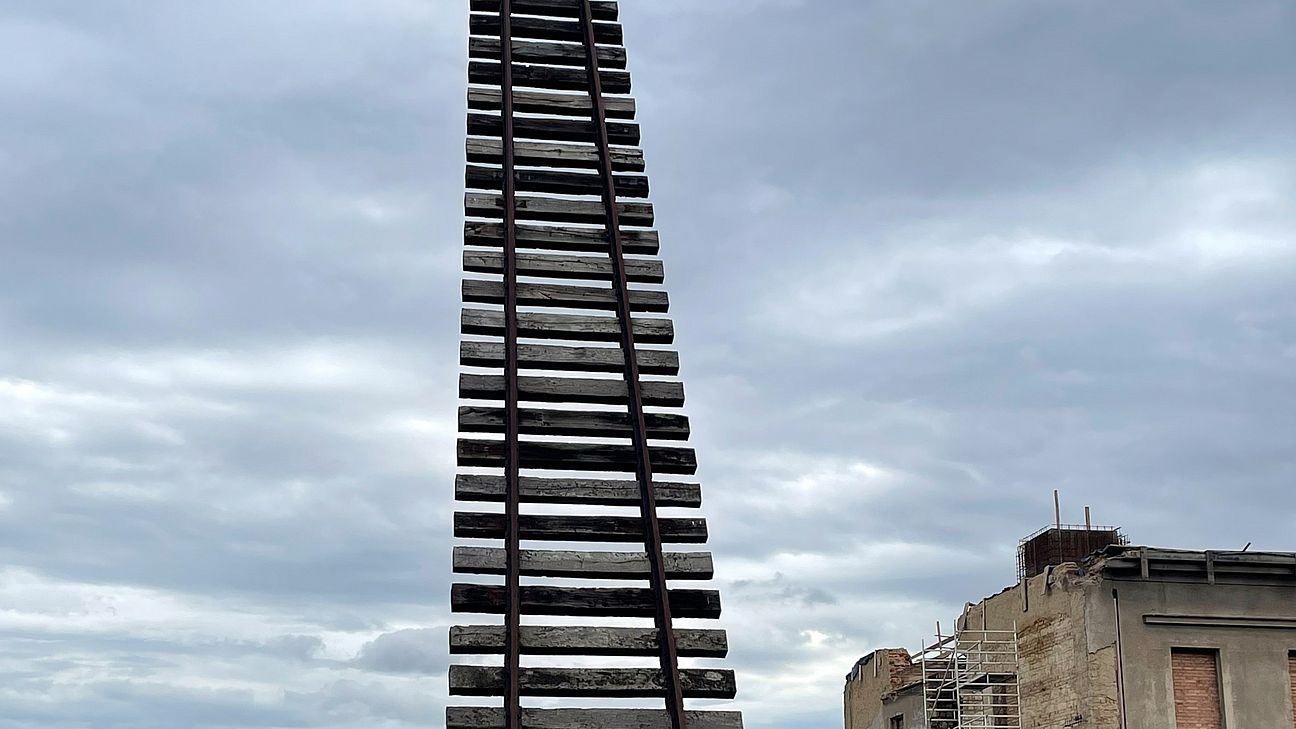 Neben der Ruine eines Gebäudes ragt eine 20 Meter hohe Skulptur aus Bahngleisen in den Himmel.