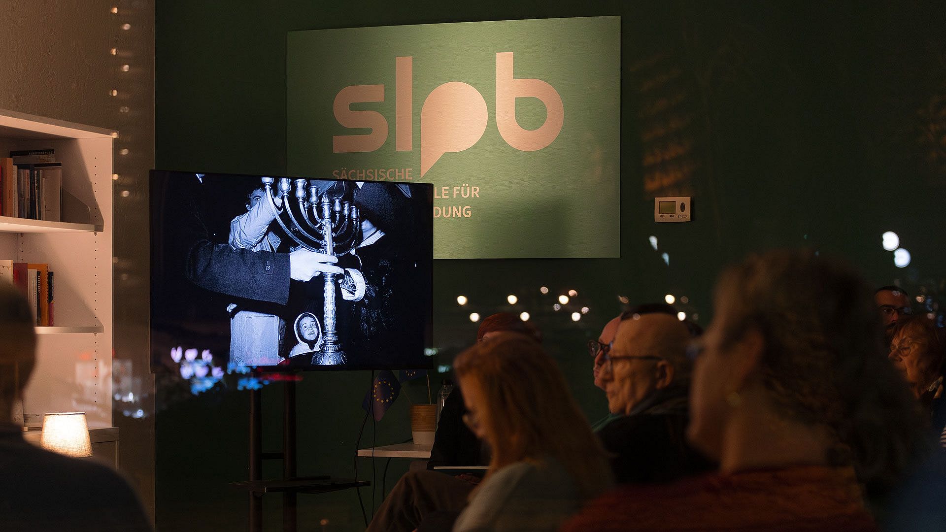 Mehrere Personen sitzen in einem dunklen Raum vor einem Bildschirm, der ein schwarz-weißes Bild zeigt von Personen die einen jüdischen Kerzenleuchter tragen, im Hintergrund ein Schild mit der Aufschrift 'slpb'.