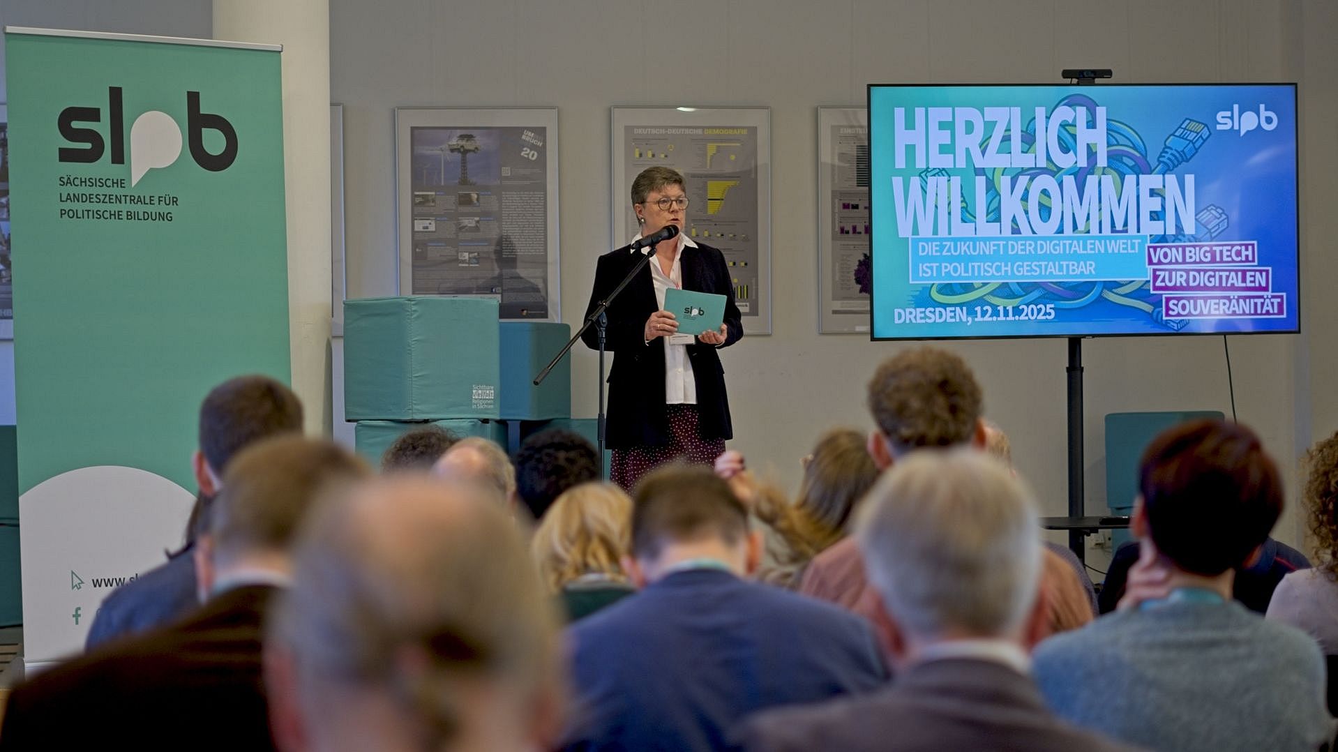 Person steht mit Mikrofon und Unterlagen vor Publikum bei der Tagung Big Tech der SLpB in Dresden. Links steht ein Banner mit dem Logo der Sächsischen Landeszentrale für politische Bildung. Rechts zeigt ein Bildschirm den Text 'Herzlich Willkommen' und Details zur Veranstaltung am 12.11.2025.