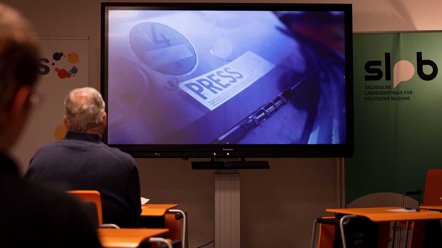 Zwei Personen sitzen vor einem Bildschirm, der eine Nahaufnahme eines Presseausweises mit dem Schriftzug 'PRESS' zeigt, im Raum mit Bannern der Sächsischen Landeszentrale für politische Bildung.