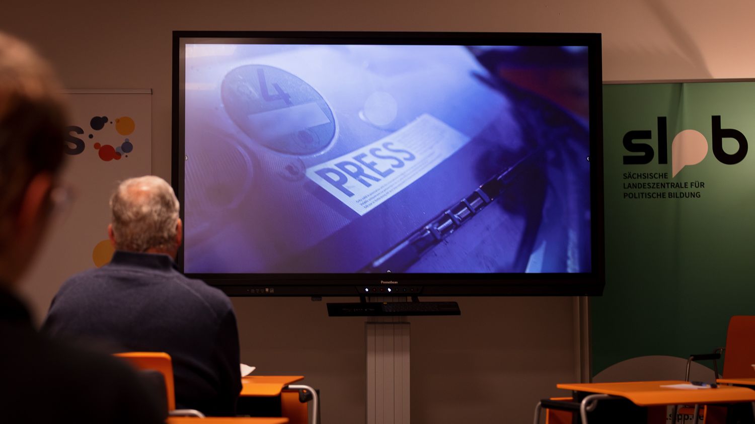 Zwei Personen sitzen vor einem Bildschirm, der eine Nahaufnahme eines Presseausweises mit dem Schriftzug 'PRESS' zeigt, im Raum mit Bannern der Sächsischen Landeszentrale für politische Bildung.