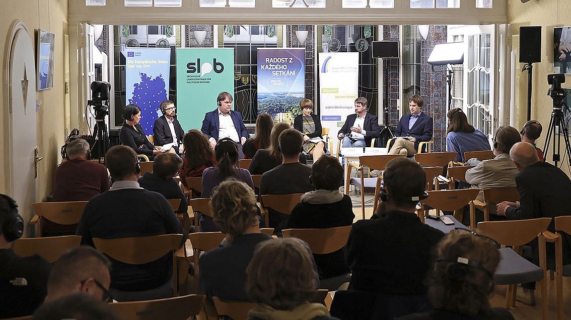 Podiumsdiskussion mit fünf Personen auf Stühlen vor Publikum in einem Raum mit hohen Fenstern und Informationsbannern im Hintergrund.
