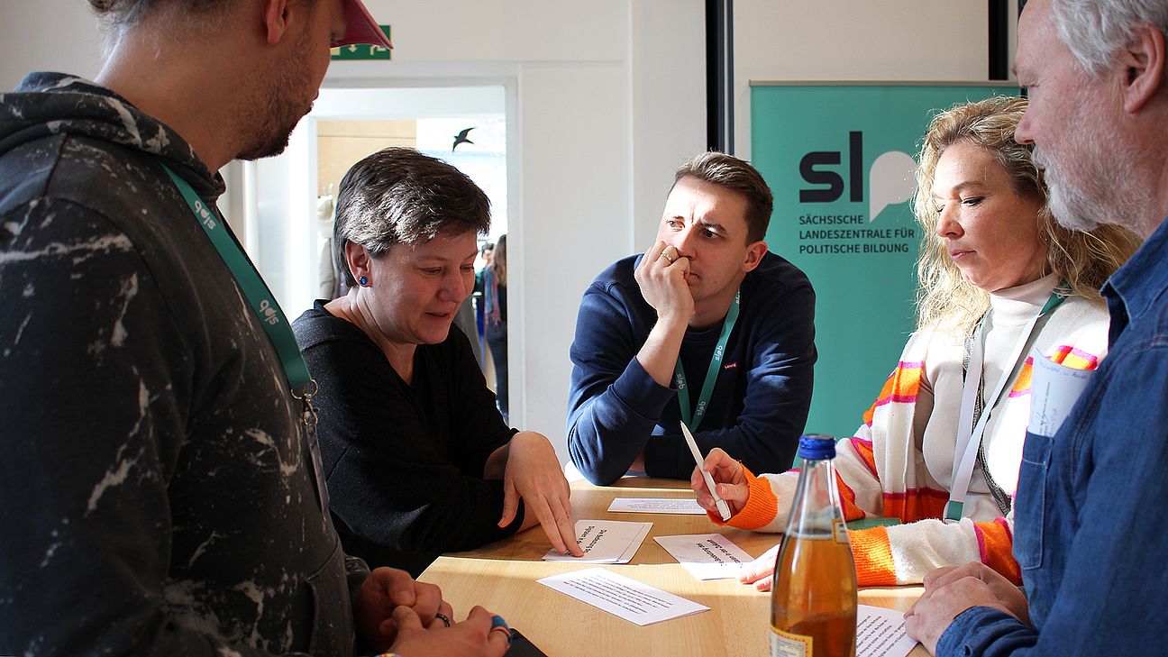 Fünf Personen sitzen und stehen um einen Tisch mit Papieren und einer Flasche, im Hintergrund ein Banner mit dem Schriftzug 'slp'.