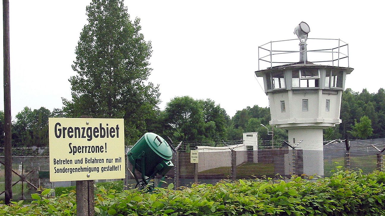Freilichtmuseum Mödlareuth Zu sehen ist ein Schild mit der Aufschrift "Grenzgebiet Sperrzone! Betreten und Befahren nur mit Sondergenehmigung gestattet!" Im Hintergrund sind ein Wachturm und ein Zaun mit Stacheldraht zu erkennen, die eine Grenze markieren.