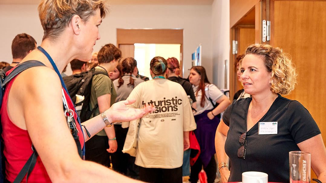 Mehrere Personen in einem Raum, eine trägt ein helles T-Shirt mit der Aufschrift 'futuristic visions', eine andere steht an einem Tisch mit Kaffeetasse und Glas Orangensaft.