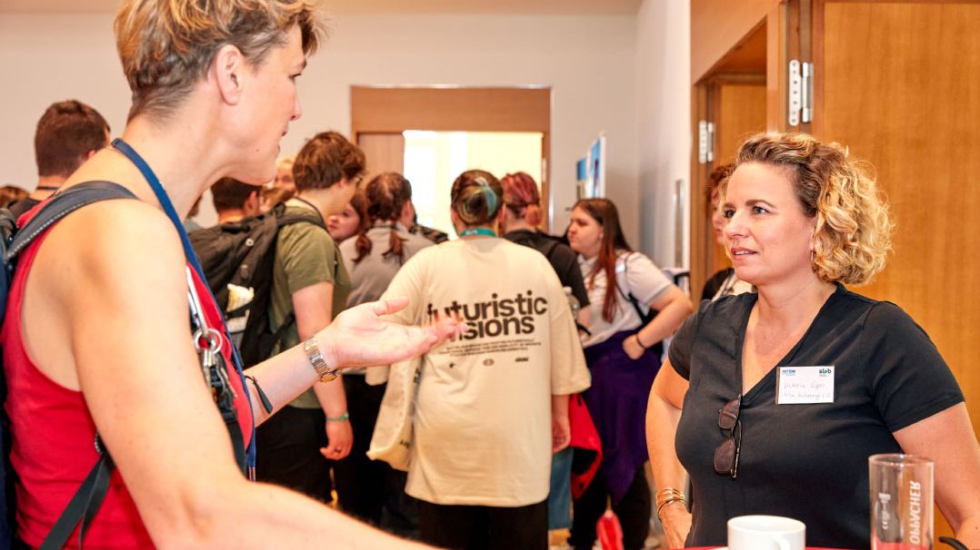 Mehrere Personen in einem Raum, eine trägt ein helles T-Shirt mit der Aufschrift 'futuristic visions', eine andere steht an einem Tisch mit Kaffeetasse und Glas Orangensaft.