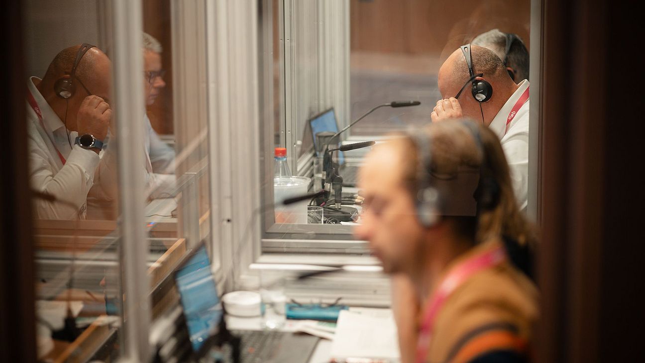 Dolmetscher in Kabinen bei der Trinationalen Tagung, mit Headsets und Mikrofonen, vor Laptops und technischer Ausrüstung sitzend.