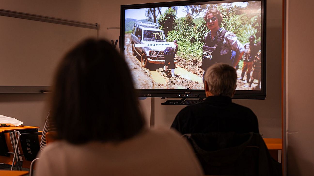 Zuschauerinnen und Zuschauer sehen eine Filmvorführung mit einer Szene, die einen Geländewagen auf einem unbefestigten Weg und Personen im Freien zeigt.
