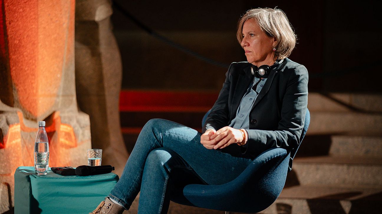 Rechtsanwältin Berenice Böhlo sitzt in einem blauen Stuhl, trägt Jeans, schwarze Jacke und Kopfhörer um den Hals, neben ihr ein Tisch mit Wasserflasche und Glas.