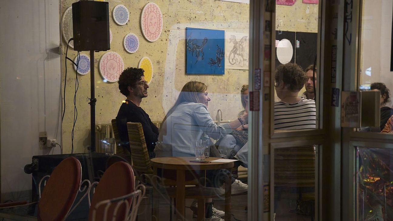 durch ein schaufenster fotografiert sieht man drei leute sitzen, die einem publikum zugewandt sind.