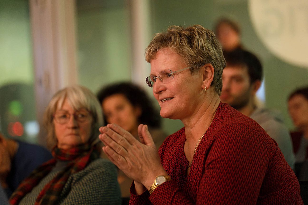 Frau mit kurzen blonden Haaren und rotem Pullover mit zusammengefalteten Händen in einem Raum mit mehreren Personen.