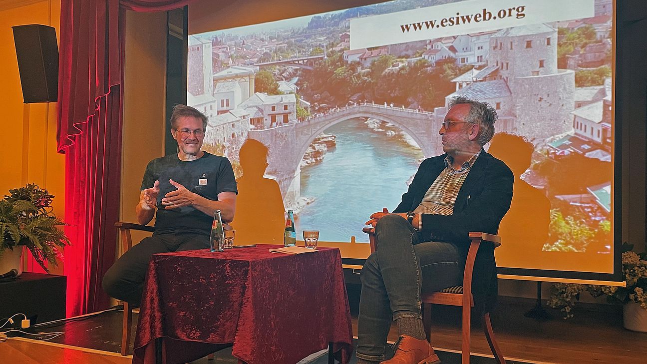 Zwei Männer sitzen auf einer Bühne vor einer Leinwand, diskutieren in einem Veranstaltungsraum mit rotem Vorhang und Tisch mit Glas und Wasserflaschen