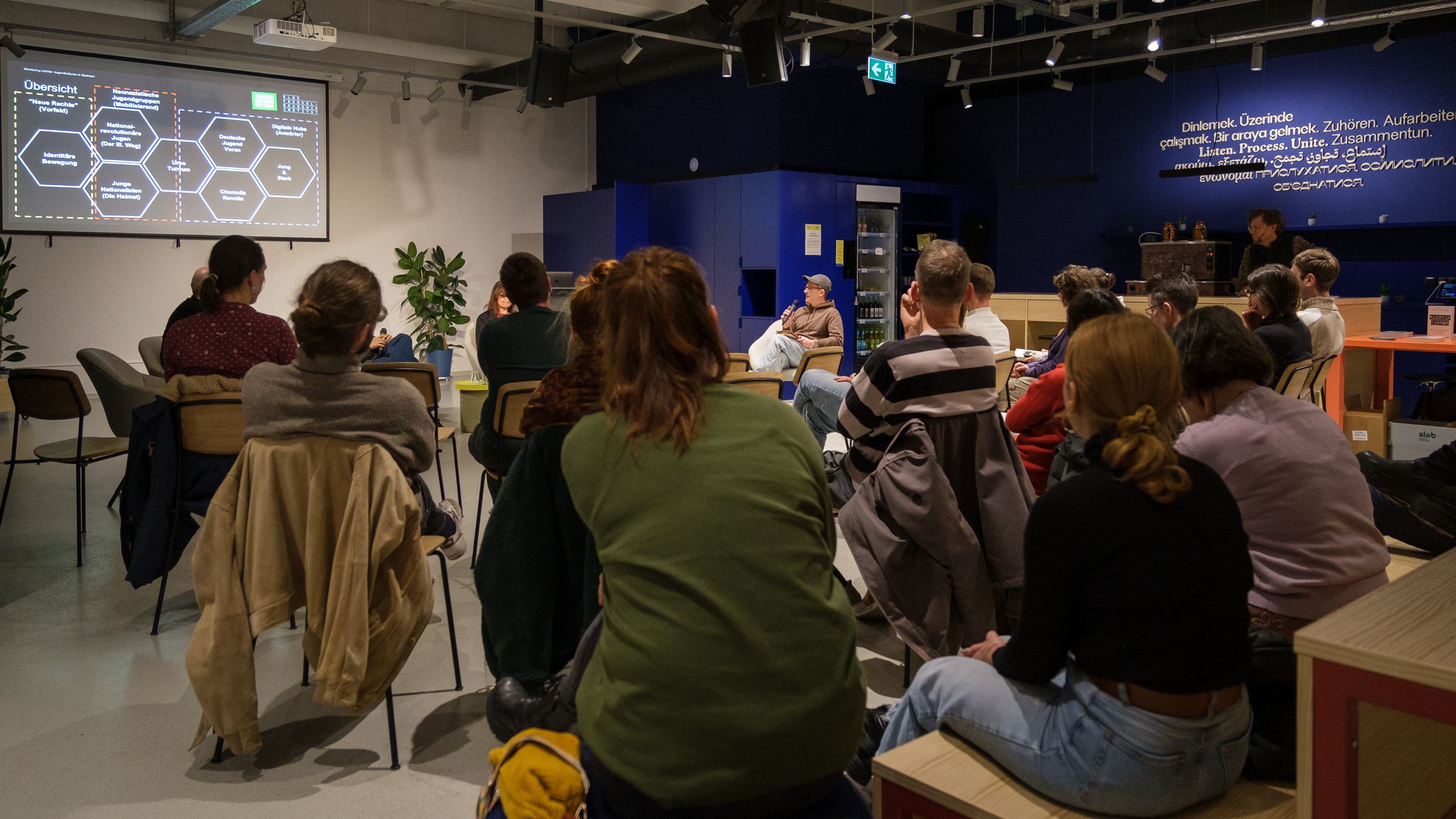 Mehrere Personen sitzen in einem Raum und schauen auf eine Präsentation mit Text und Diagrammen an der Wand.