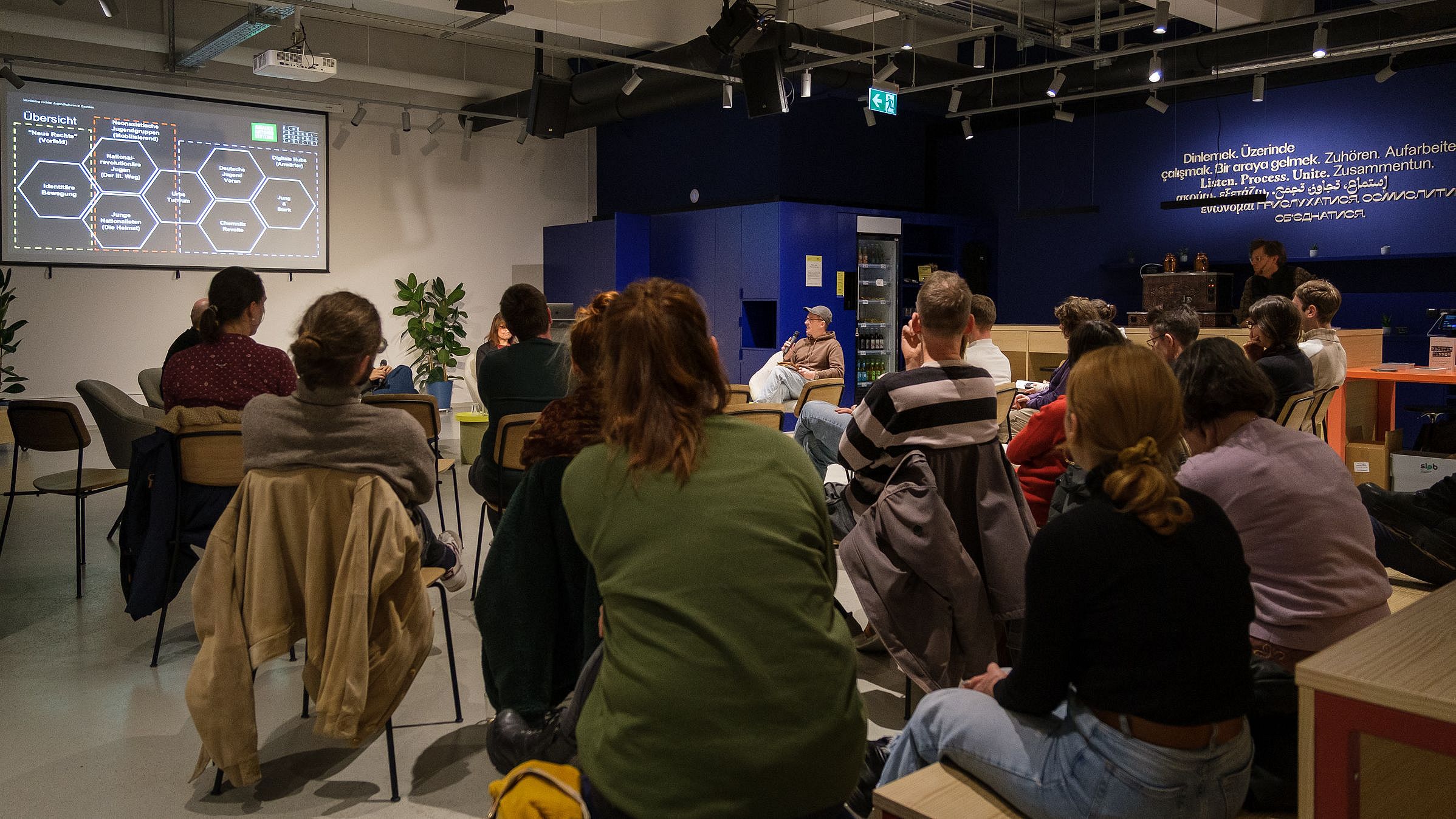 Mehrere Personen sitzen in einem Raum und schauen auf eine Präsentation mit Text und Diagrammen an der Wand.