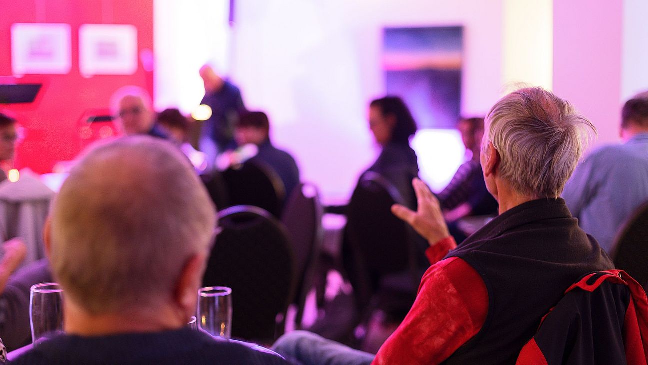 Blick in den Veranstaltungsraum während der Publikumsdiskussion. Eine Besucherin mit kurzen grauen Haaren hebt ihre Hand und gestikuliert. Weitere Gäste sitzen an Tischen und hören zu.