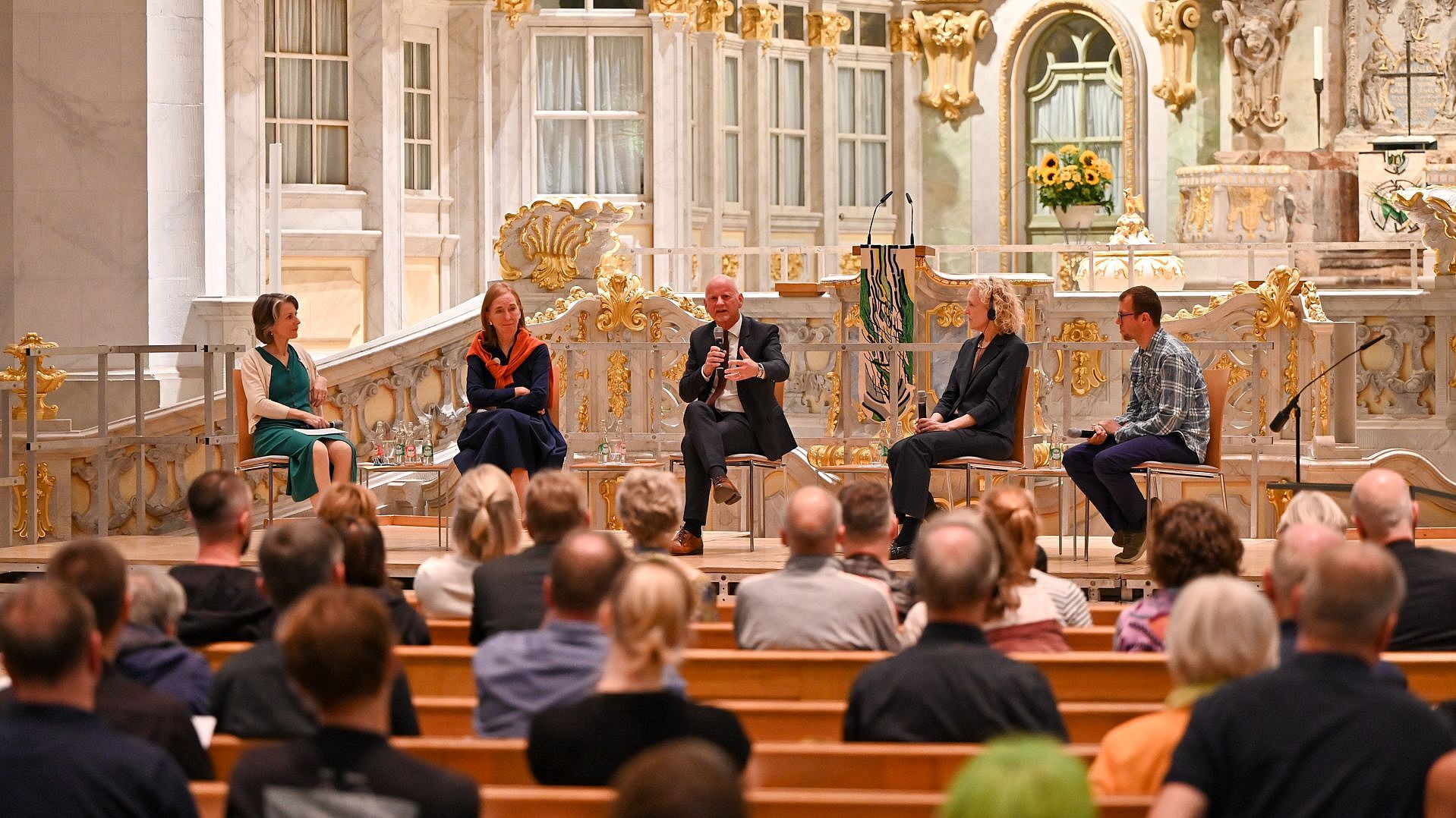 Fünf Personen sitzen auf Stühlen vor einer barocken Altarwand in der Frauenkirche Dresden und sprechen vor einem Publikum auf Holzbänken.