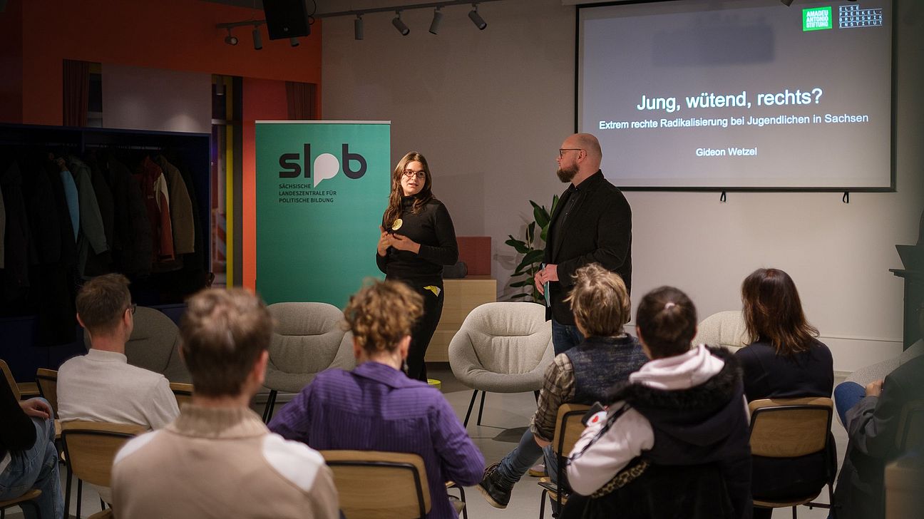 Zwei Personen stehen vor Publikum in einem Raum mit Stühlen, eine Leinwand zeigt den Titel 'Jung, wütend, rechts? Extrem rechte Radikalisierung bei Jugendlichen in Sachsen'.