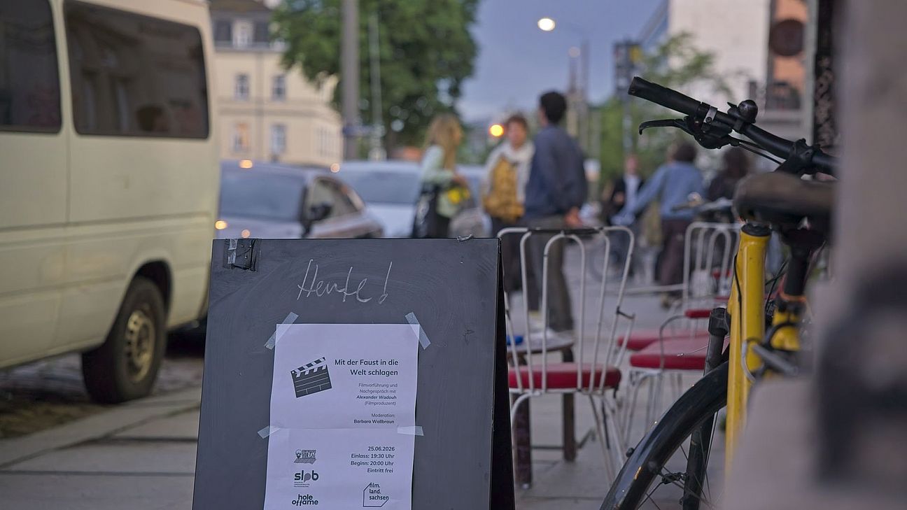 eine infotafel steht auf dem fussweg. dahinter einige sitzgelegenheiten. links daneben geparkte autos und im hintergrund eine unscharfe gruppe menschen