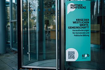 Auf diesem Bild ist ein Poster oder eine Plakatwand zu sehen, die an einer Glaswand oder einem Eingang eines Gebäudes angebracht ist. Der Text auf dem Plakat lautet:      Im oberen Teil des Plakats in großer Schrift "PARTNERKONFERENZ".     Darunter steht in größerer, fetter Schrift "KRISE DER WESTLICHEN WERTE-GEMEINSCHAFT?".     In einer kleineren Schrift darunter steht "Herausforderungen für Demokratie und Politische Bildung".     Am unteren Rand des Plakats befindet sich ein QR-Code, der vermutlich zur Webseite oder zum Programm der Konferenz führt.     Unter dem QR-Code ist das Logo oder die Abkürzung "SLpB" zu sehen, was für die "Sächsische Landeszentrale für politische Bildung" steht.  Das Design des Plakats ist schlicht und nutzt eine ähnliche Farbgebung wie das zuvor gesehene Bild der mobilen Webseite oder App – hauptsächlich in Grüntönen. Es scheint Werbung für eine Veranstaltung oder Konferenz zu sein, die sich mit aktuellen gesellschaftlichen und politischen Fragen beschäftigt.