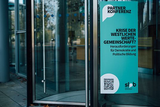 Auf diesem Bild ist ein Poster oder eine Plakatwand zu sehen, die an einer Glaswand oder einem Eingang eines Gebäudes angebracht ist. Der Text auf dem Plakat lautet:      Im oberen Teil des Plakats in großer Schrift "PARTNERKONFERENZ".     Darunter steht in größerer, fetter Schrift "KRISE DER WESTLICHEN WERTE-GEMEINSCHAFT?".     In einer kleineren Schrift darunter steht "Herausforderungen für Demokratie und Politische Bildung".     Am unteren Rand des Plakats befindet sich ein QR-Code, der vermutlich zur Webseite oder zum Programm der Konferenz führt.     Unter dem QR-Code ist das Logo oder die Abkürzung "SLpB" zu sehen, was für die "Sächsische Landeszentrale für politische Bildung" steht.  Das Design des Plakats ist schlicht und nutzt eine ähnliche Farbgebung wie das zuvor gesehene Bild der mobilen Webseite oder App – hauptsächlich in Grüntönen. Es scheint Werbung für eine Veranstaltung oder Konferenz zu sein, die sich mit aktuellen gesellschaftlichen und politischen Fragen beschäftigt.