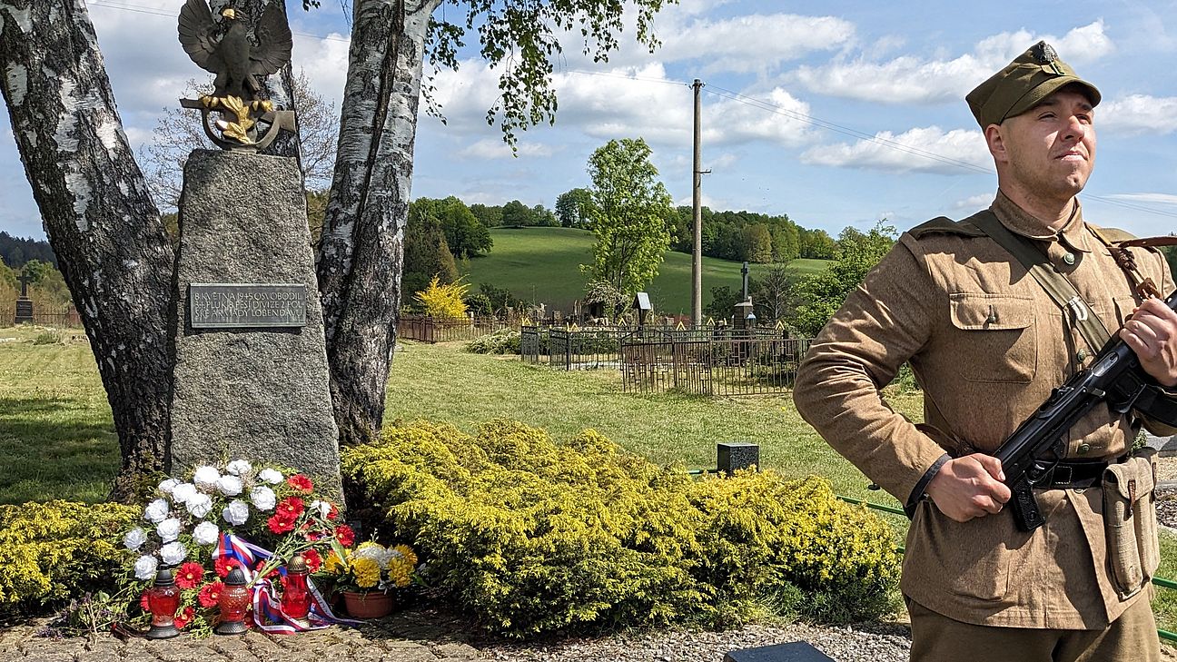 auf der rechten Seite steht eine Person in Uniform der damaligen 2. Polnischen Armee mit einer Waffe in der Hand. Links im Mittelpunkt ist ein Mahnmal zu sehen, vor dem Blumen stehen. Im Hintergrund sind Gräber zu sehen.