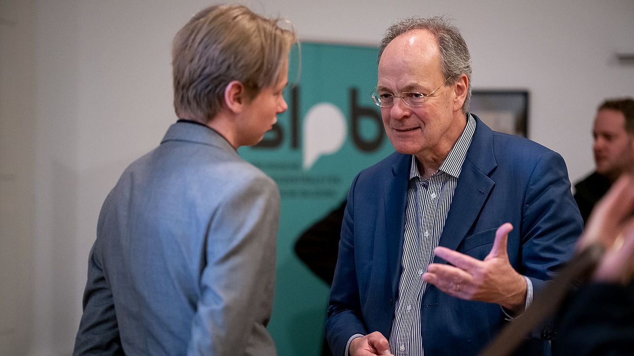 Zwei Personen im Gespräch, eine trägt einen grauen Anzug, die andere einen blauen Anzug, im Hintergrund das grüne Banner mit Logo der SLpB.
