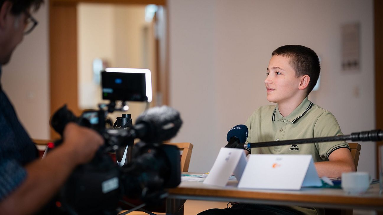 Rechts sitzt eine junge Person vor einem Mikrofon und wird von einem Kameralicht angestrahlt, links steht eine unscharfe Person mit einer Kamera und filmt.