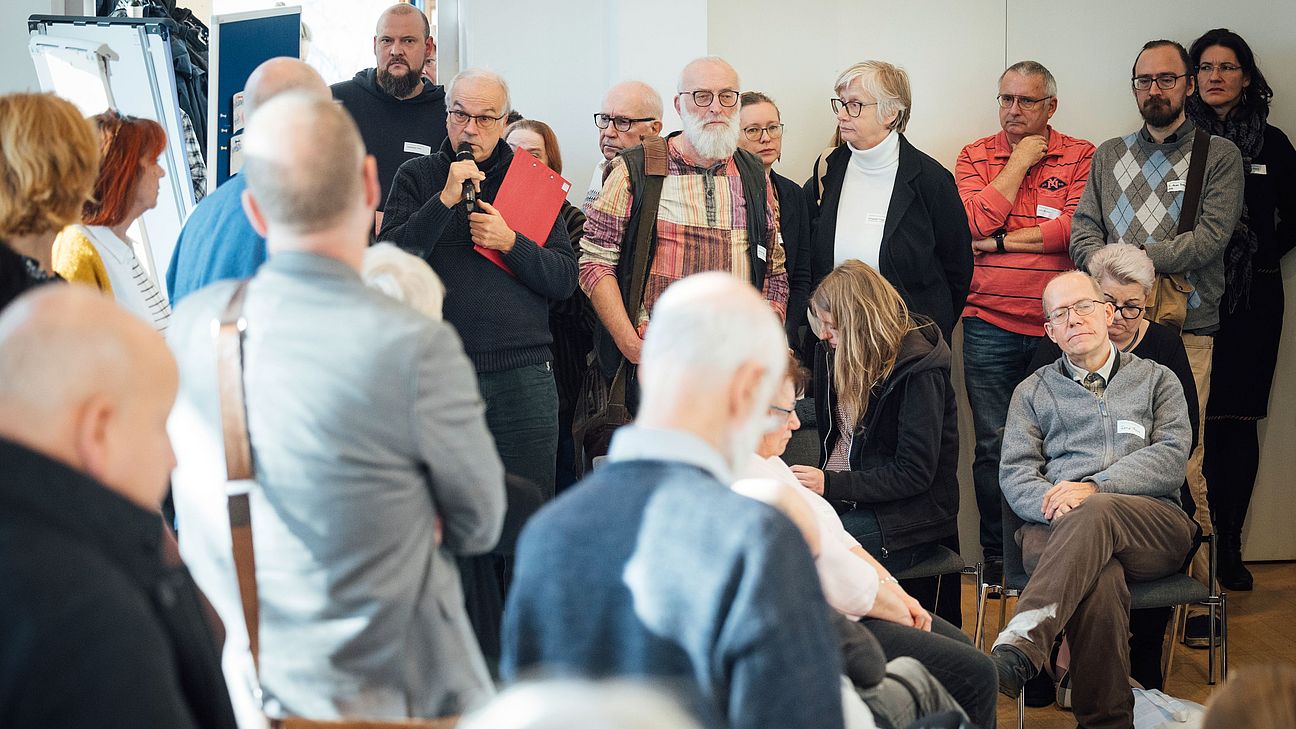 Viele Leute stehen und sitzen gedrängt bei einer Tagung um eine Moderationswand. Eine ältere Person hat ein Mikrofon in der Hand.