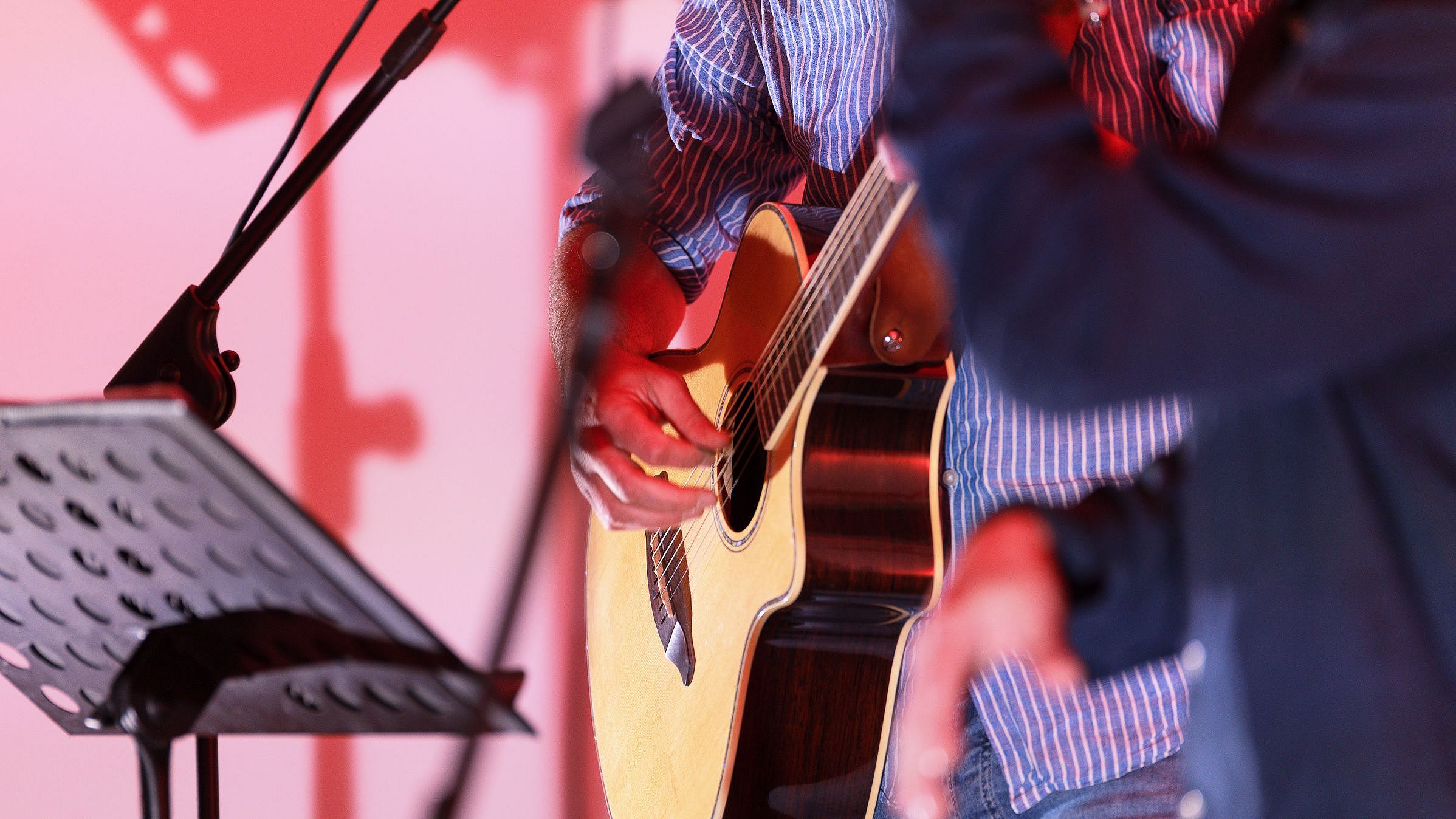 Nahaufnahme einer akustischen Gitarre auf einer rot beleuchteten Bühne. Im Vordergrund ist eine weitere Person unscharf zu sehen.