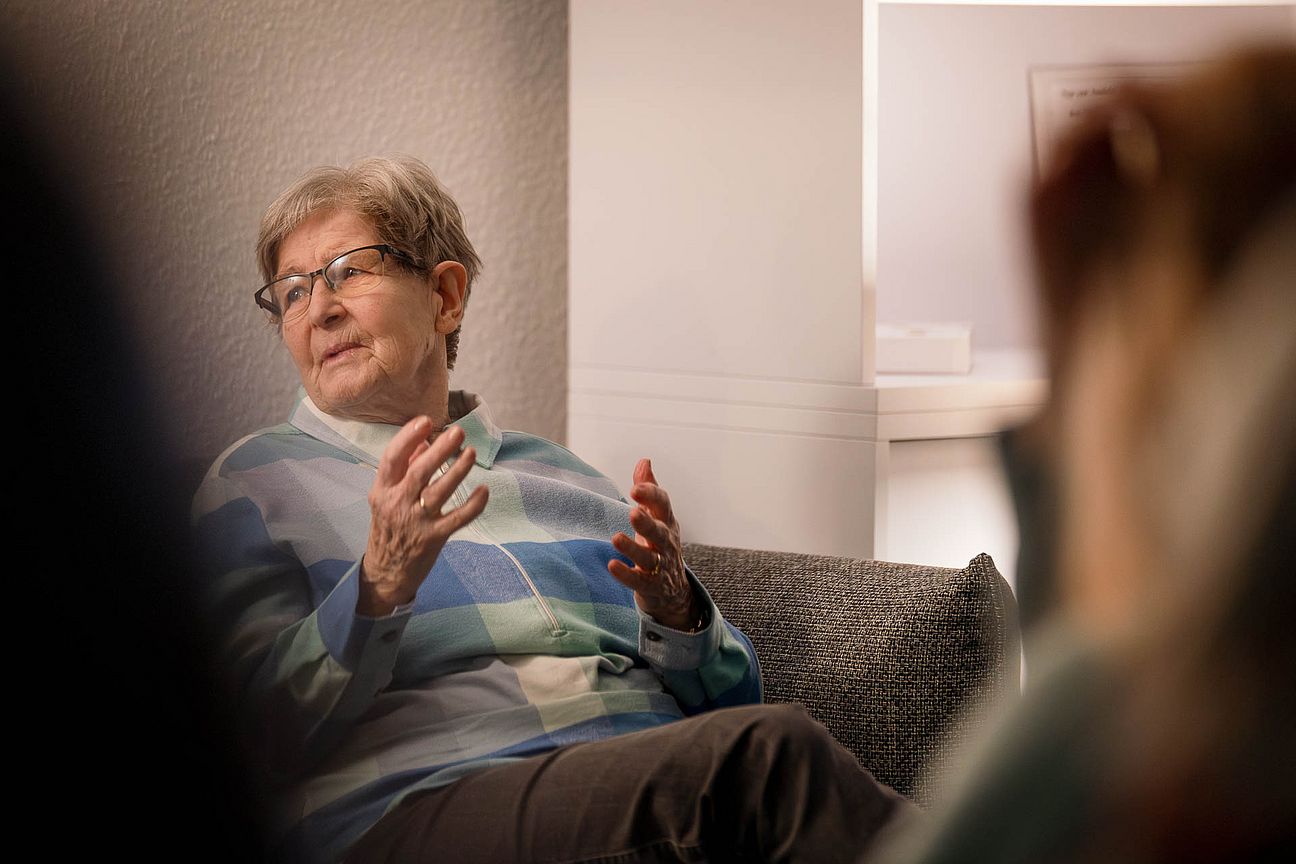 Ältere Person mit Brille und gestreiftem Pullover sitzt auf einem Sofa und gestikuliert mit den Händen.