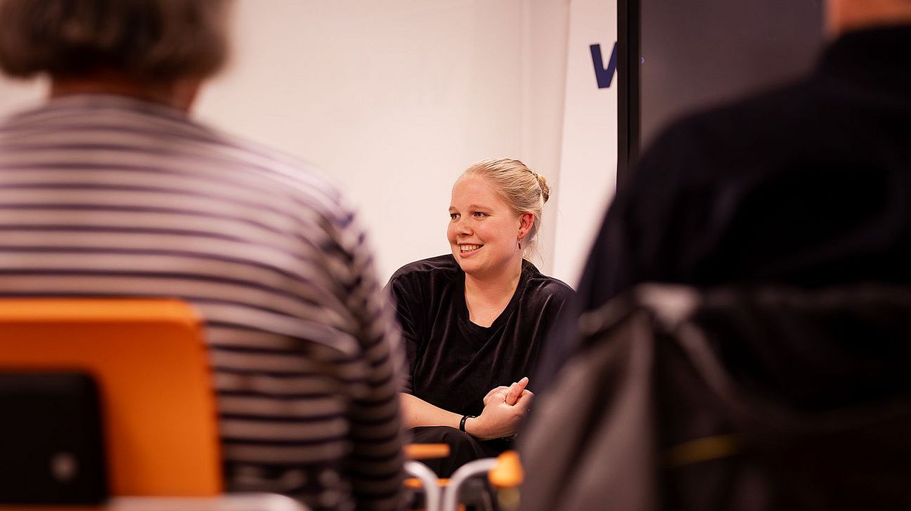 Person mit blondem Haar und schwarzem Oberteil sitzt vor Publikum in einem Raum mit weißen Wänden und hölzernenStühlen.