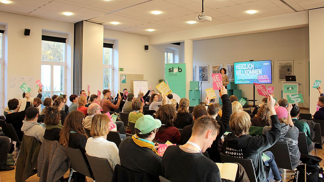 Mehrere Personen sitzen in einem hellen Seminarraum und heben bunte Schilder mit Texten hoch. Im Hintergrund ein Bildschirm mit der Aufschrift 'HERZLICH WILLKOMMEN'.